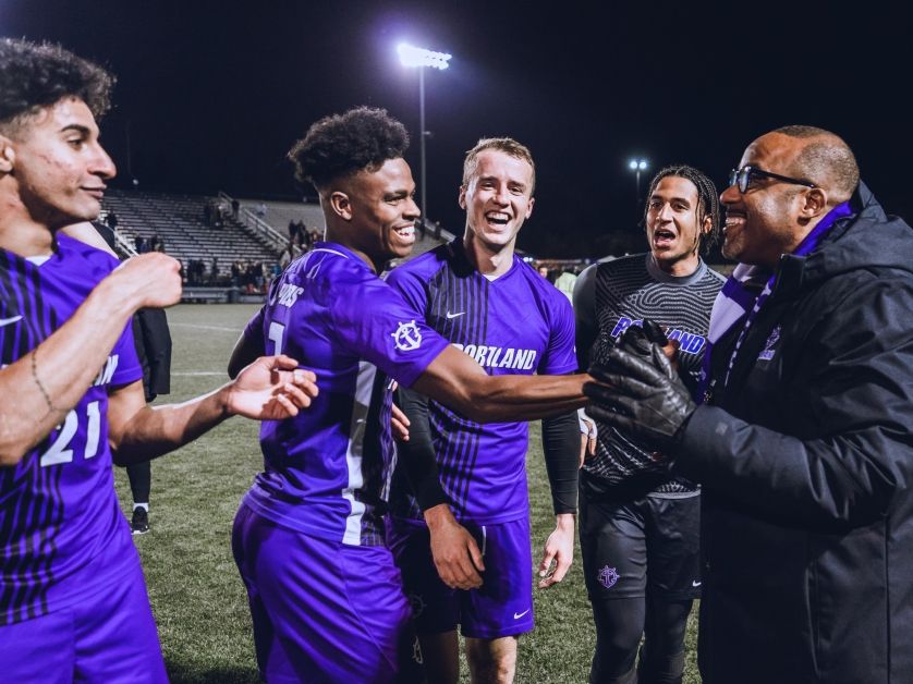 A group of men's soccer players shake hands with UP President Kelly on the soccer pitch during a night game.